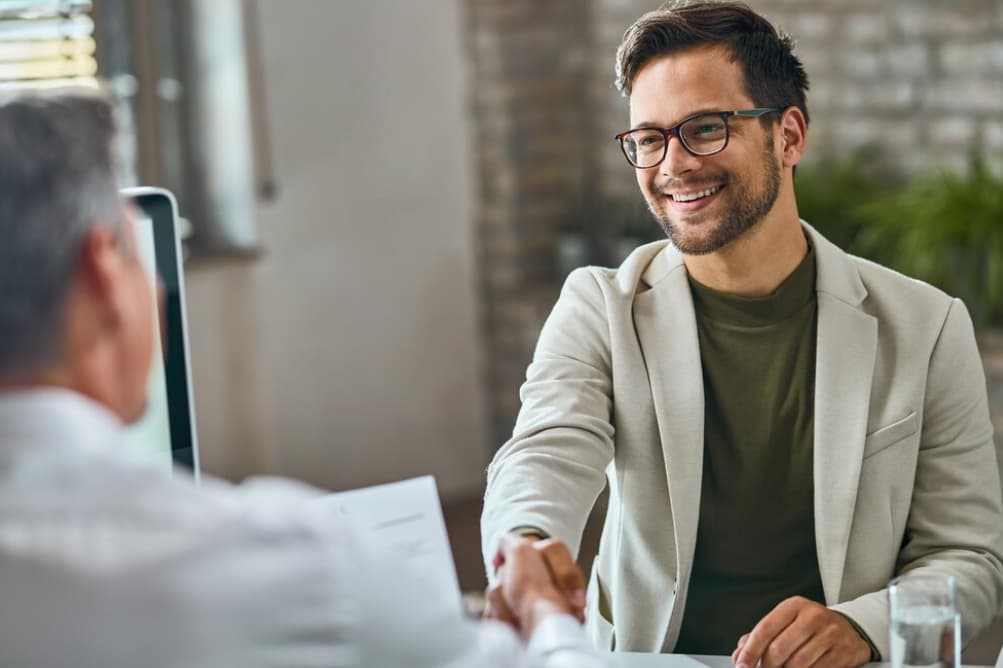 A smiling man in glasses shaking hands across a desk