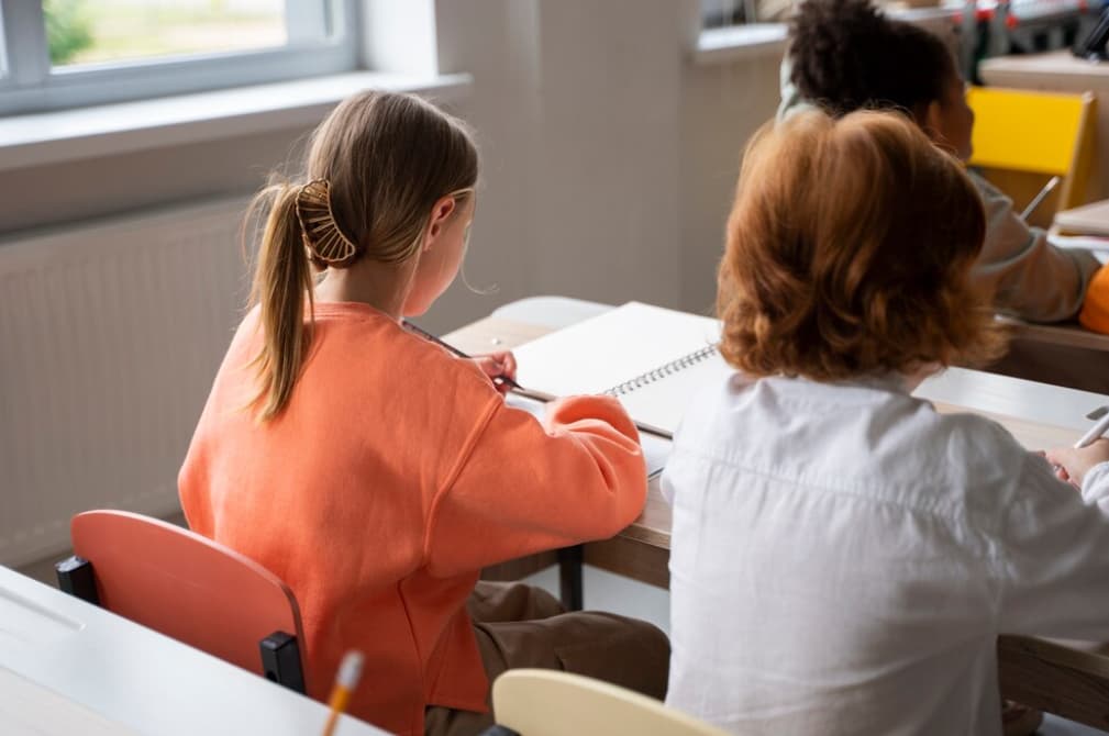 A student takes notes while facing a teacher in a bright classroom